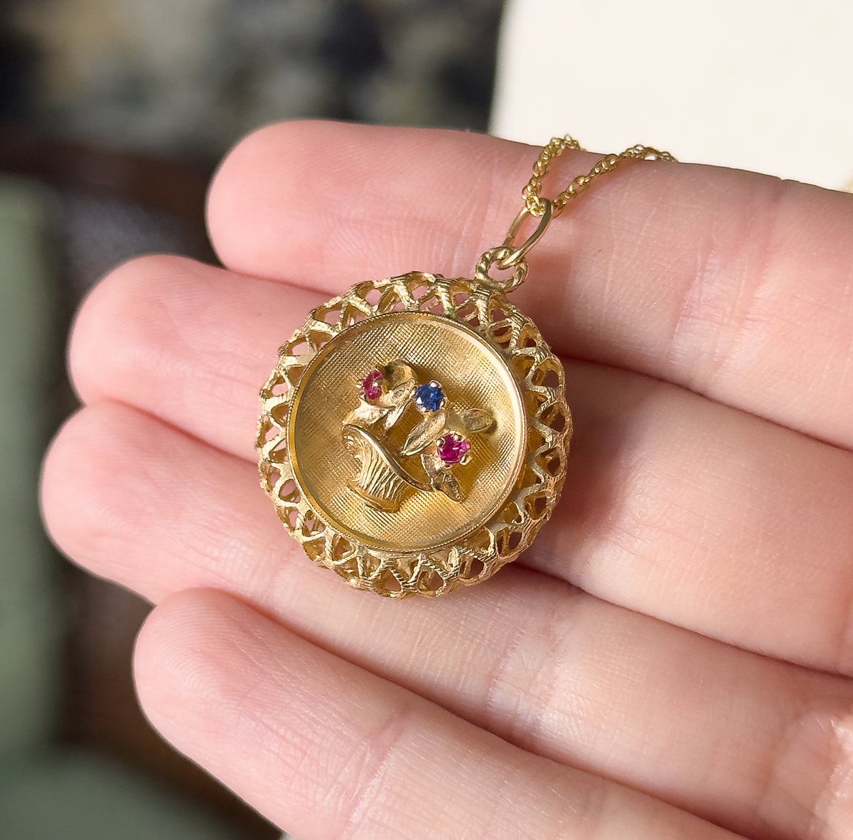 A 14K yellow gold flower basket pendant necklace, shown resting on fingertips, its lattice openwork gallery framing a textured basket set with two red and one blue small stones.
