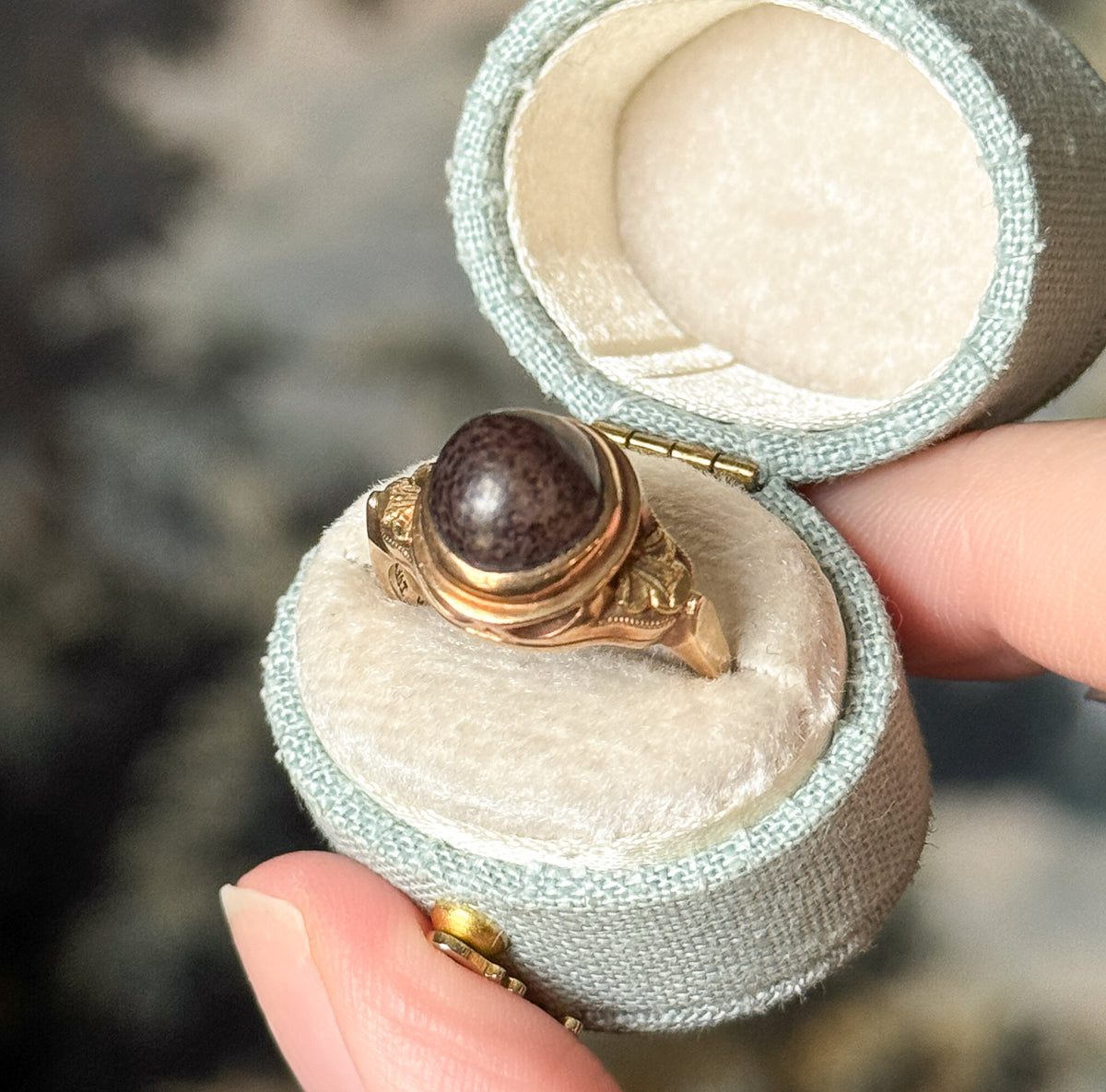 Art Deco brown stone cabochon ring in 10K rose gold, shown in a small pale mint ring box held between finger and thumb, the round stone set in a bezel above decorated shoulders.