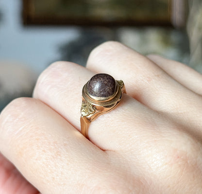 Shown on the hand from above at an angle, the ring sitting between two fingers so its scale is clear, with the domed brown cabochon and its bezel facing the viewer.