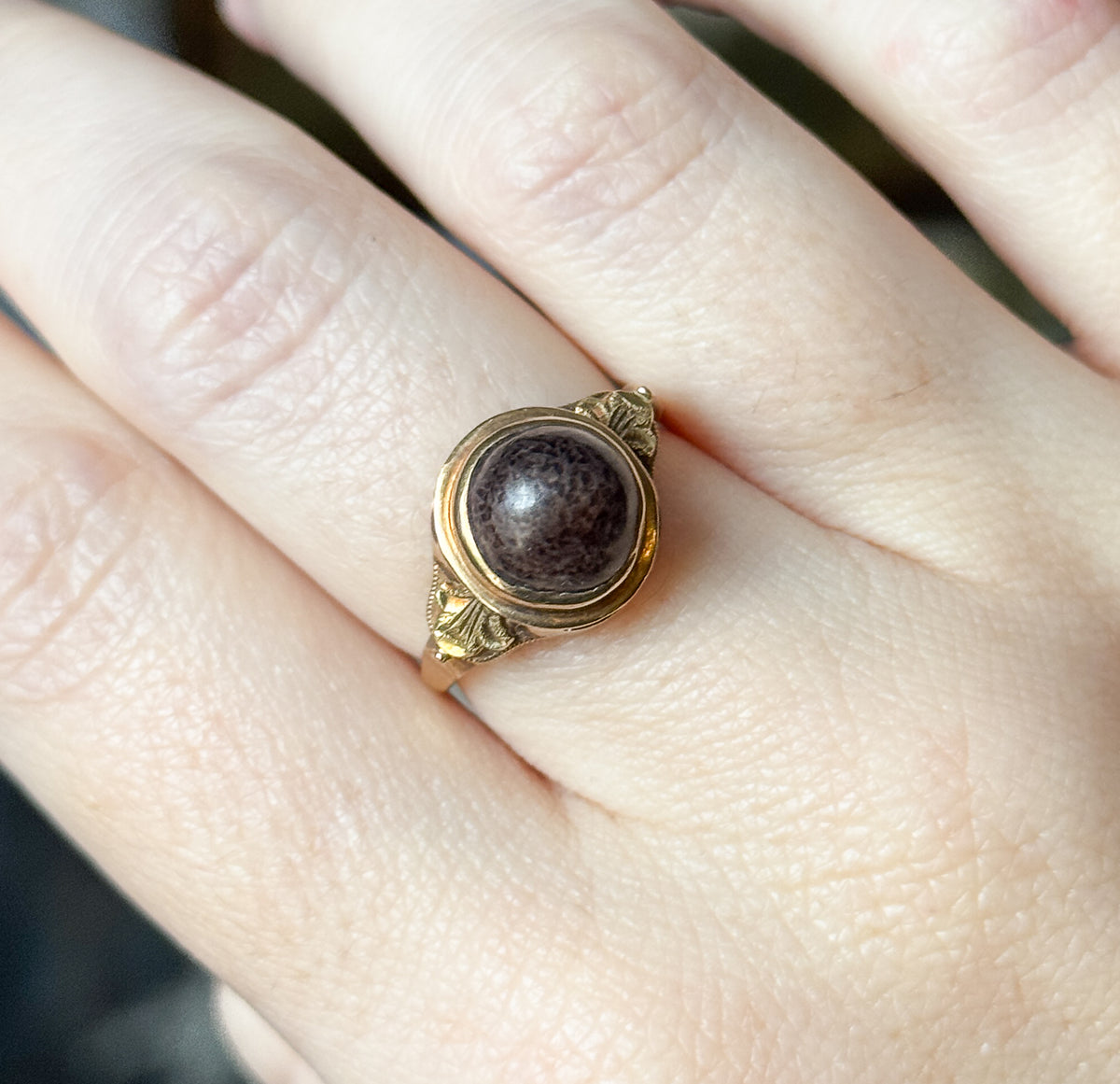 Shown on the hand in close crop, the bezel and its rose gold rim filling the frame so the speckled brown cabochon and leafy shoulder details read clearly against the skin.