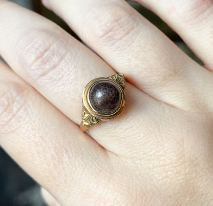Shown on the hand in close crop, the bezel and its rose gold rim filling the frame so the speckled brown cabochon and leafy shoulder details read clearly against the skin.