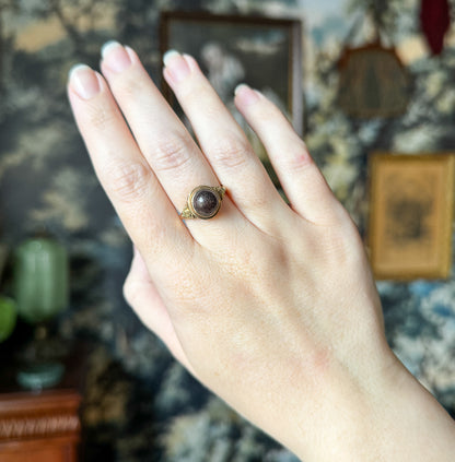 Shown on the hand held upright and open against a patterned interior, the whole hand giving scale as the small brown cabochon and its bezel sit low on the ring finger.