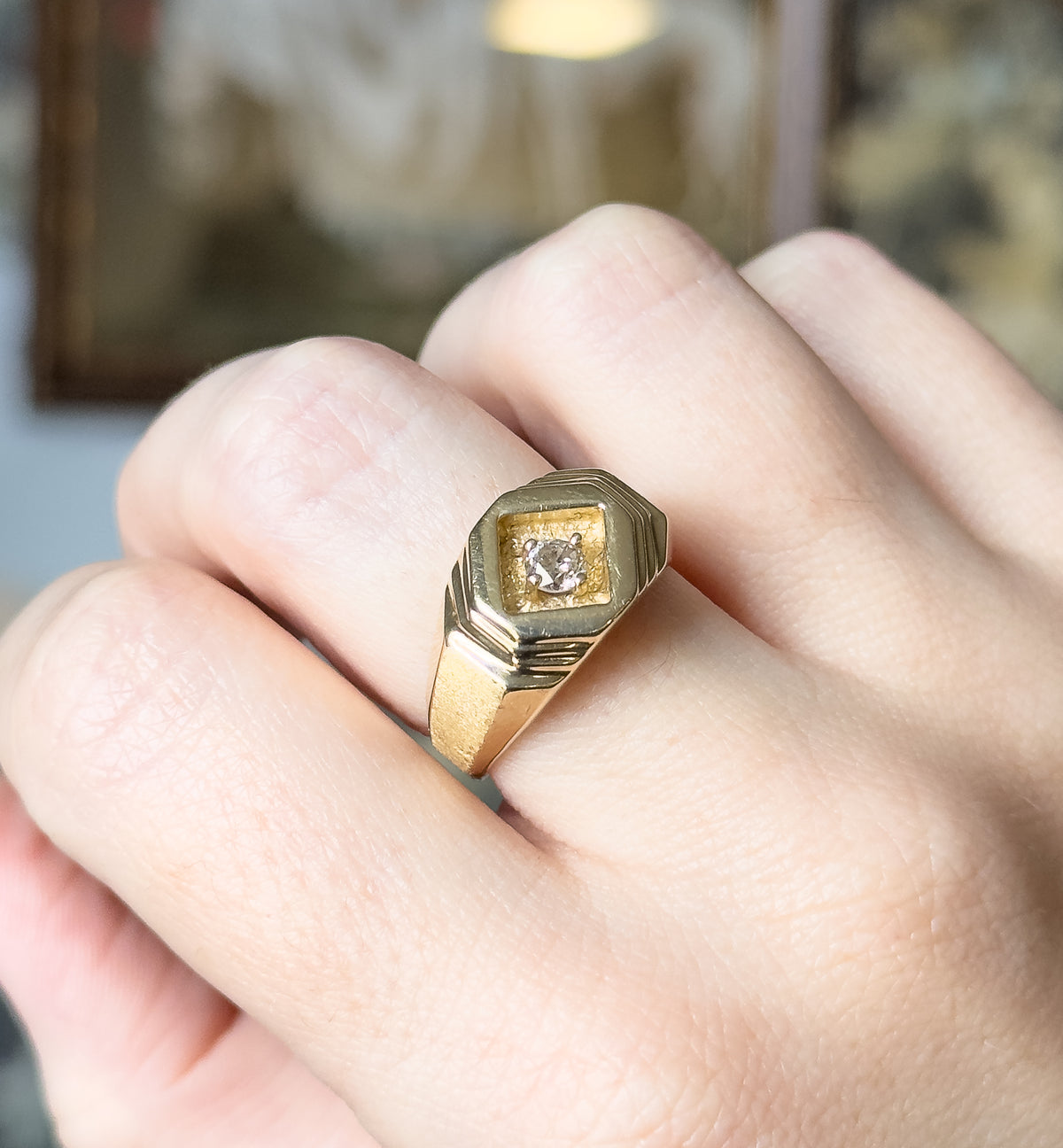 A geometric 14K yellow gold ring with a round diamond prong-set in a recessed diamond-shaped face, shown on a finger with the stepped shoulders and tapering shank visible.