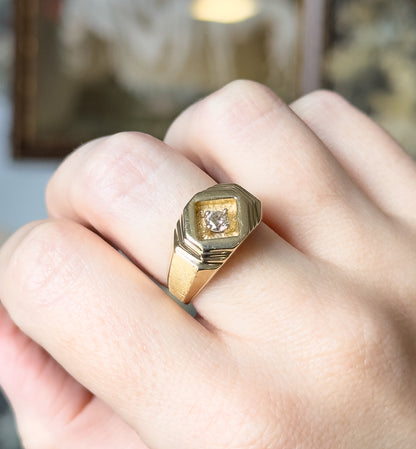 A geometric 14K yellow gold ring with a round diamond prong-set in a recessed diamond-shaped face, shown on a finger with the stepped shoulders and tapering shank visible.