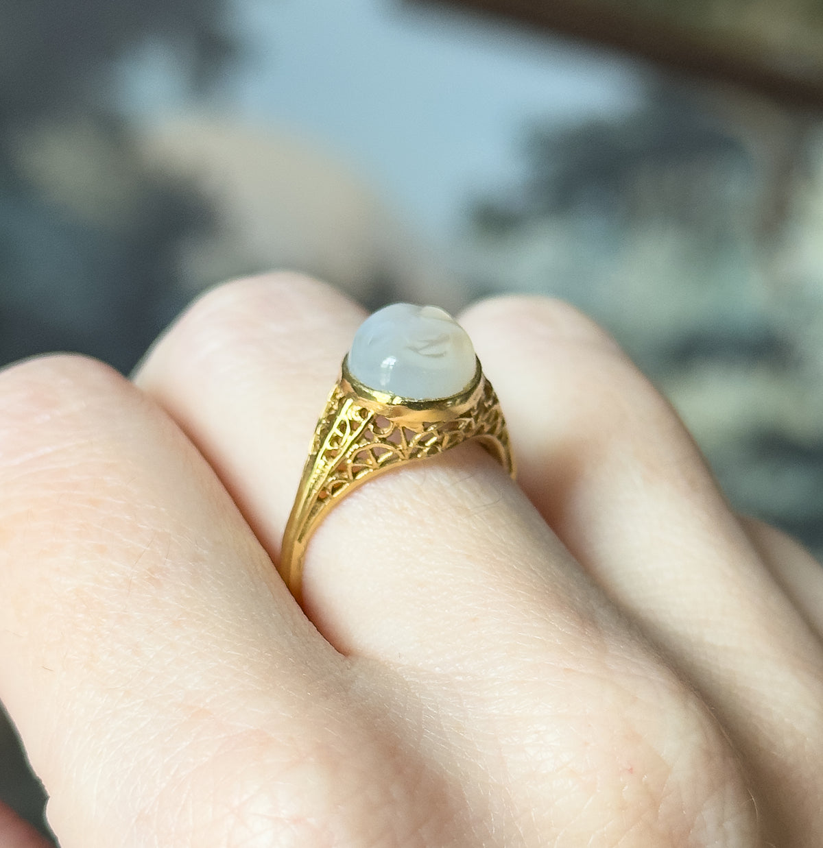 Closer three-quarter view shown on a finger, the pale carved moonstone cabochon catching light in its bezel above openwork filigree shoulders that narrow into the shank.