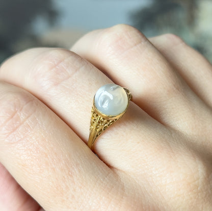Head-on view shown on a finger, the carved lunar face on the round moonstone cabochon turned fully toward the camera above the filigree shoulders.
