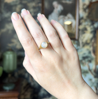 Back of the hand raised against a dark painted screen, the ring shown on the third finger so its carved moonstone face reads small and pale beside the neighbouring knuckles.