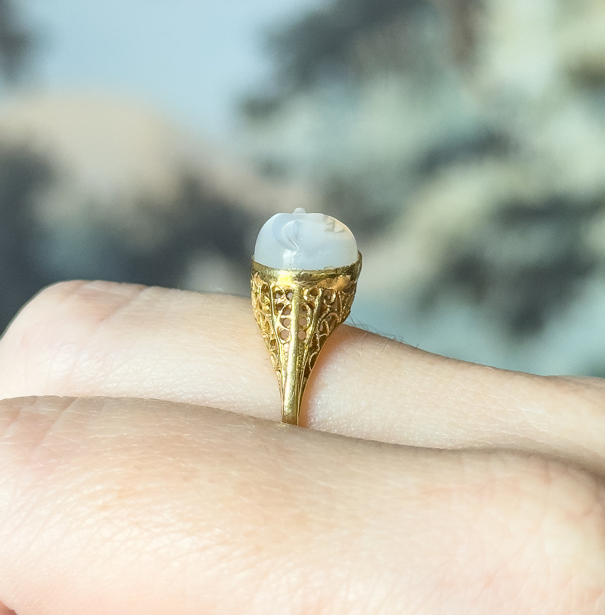 Art Deco carved moonstone Man on the Moon ring in 18K yellow gold, shown on a finger in profile so the filigree gallery and tapering shank are visible against a blurred outdoor background.