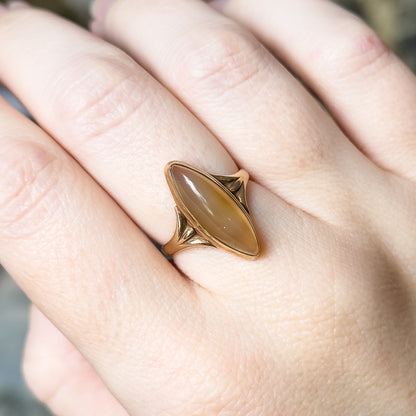 Close overhead view on the finger, the navette bezel seen straight on with the pointed rose gold shoulders flaring to either side of the golden brown cabochon.