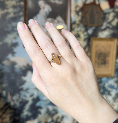 Full view of the back of the hand against a blurred framed-picture backdrop, the small navette ring on the ring finger giving a clear sense of its scale.