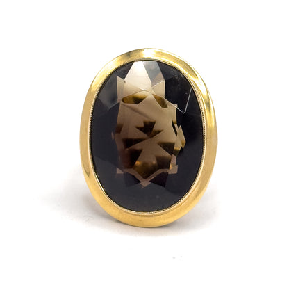 Face-on and filling the frame, the oval smoky quartz sits deep in its smooth yellow gold bezel, its faceted crown paling to honey at the centre against the dark brown depths.