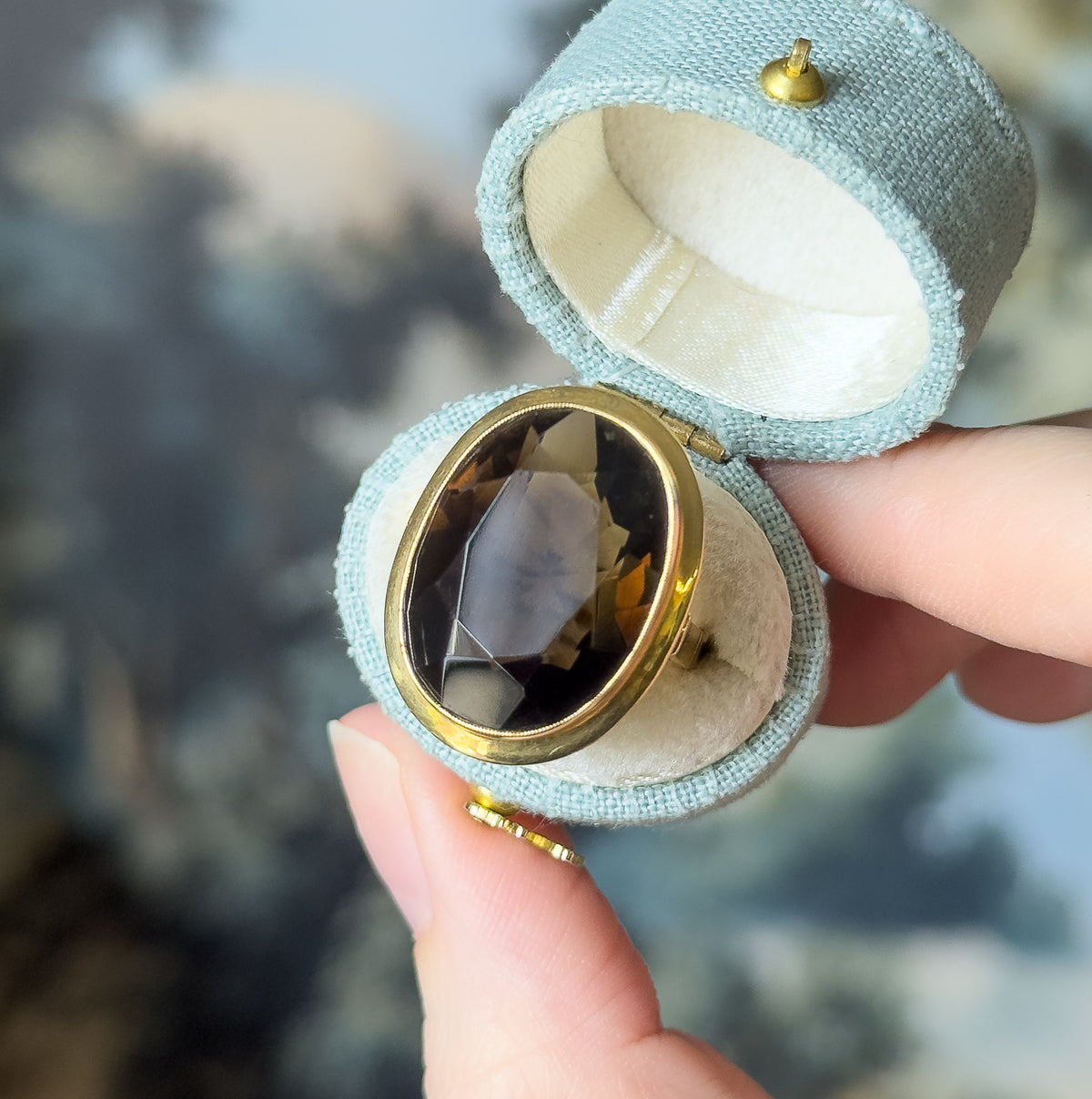 A midcentury smoky quartz cocktail ring in 14K yellow gold, its large oval bezel-set stone facing up, shown seated in a small pale blue fabric ring box held between finger and thumb.
