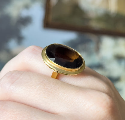 Shown on a finger at a three-quarter angle, the deep brown oval filling the width of the finger, its scalloped yellow gold bezel and narrow shank lit against a blurred background.