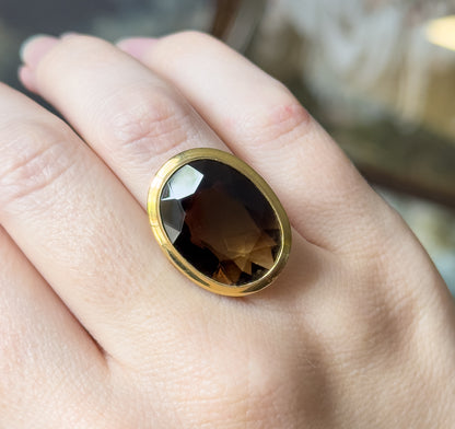 Shown on a finger from directly above, the oval stone's broad facets catching light across its chocolate-brown surface within the smooth yellow gold bezel.