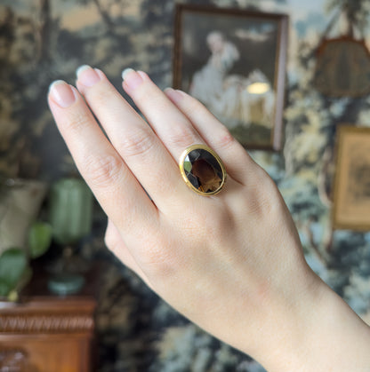 Shown on the ring finger of a hand held up against a framed picture and patterned wall, the broad oval face spanning the finger and giving a clear sense of its size.
