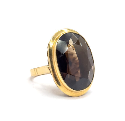 Upright on white with the oval face turned three-quarters toward the viewer, the raised yellow gold bezel rim curving around the smoky quartz while the slim shank drops away behind.