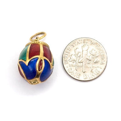 An enamel egg charm in 14K yellow gold, shown upright beside a dime for scale, its blue, red and green panels divided by gold ribs with a rope-textured collar and round bail on top.