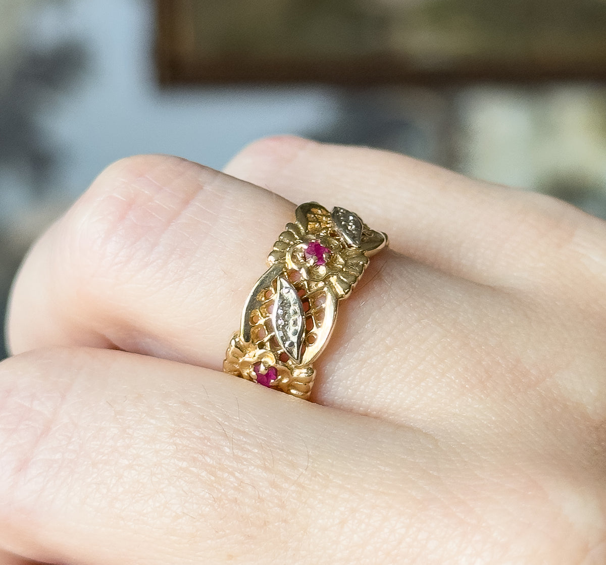 Ruby lattice and leaf ring in 14K two-tone gold, shown on a finger in close-up, its wide band showing white and yellow gold leaves with small prong-set deep pinkish red stones.