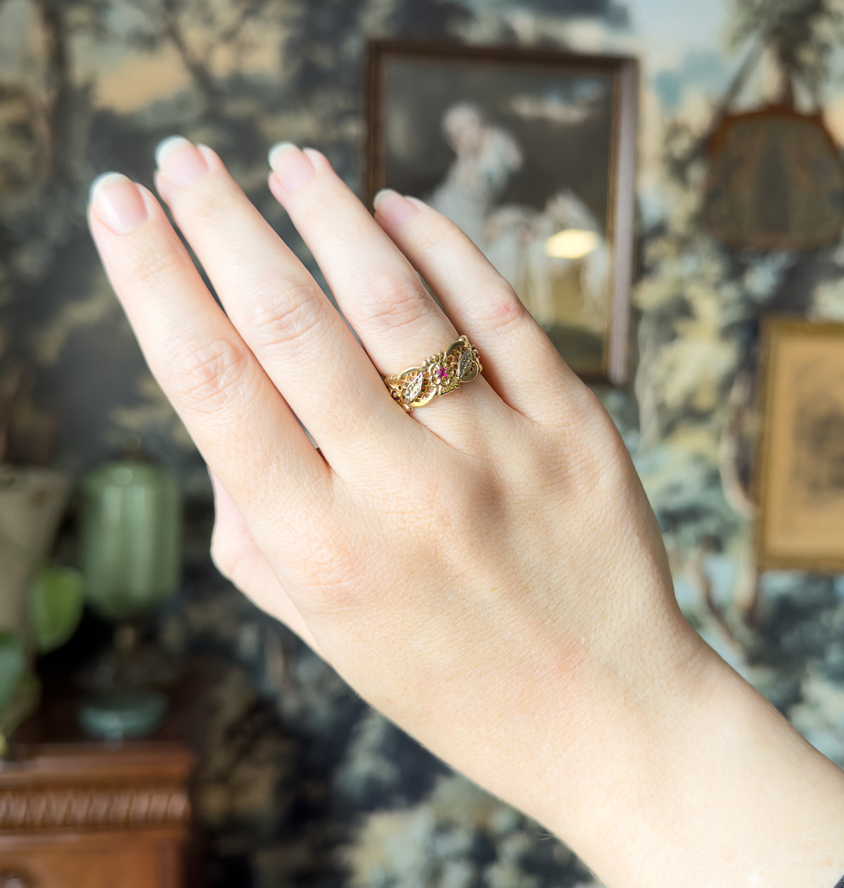 The hand is held up against a soft painted backdrop, the wide two-tone band small on the ring finger, giving a sense of its scale against the whole hand.
