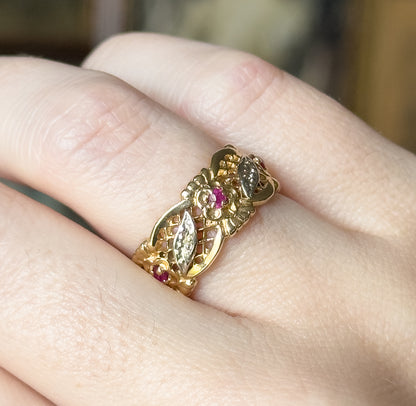 Close overhead view across the finger, showing the band's face in detail: a central flower with a deep pinkish red stone at its heart, flanked by pointed leaf panels set with tiny pale stones.
