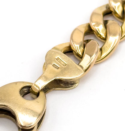 A tight macro of the flat shield-shaped end link bearing two small rectangular stamps, with the chunky yellow gold curb links curving away beyond it.