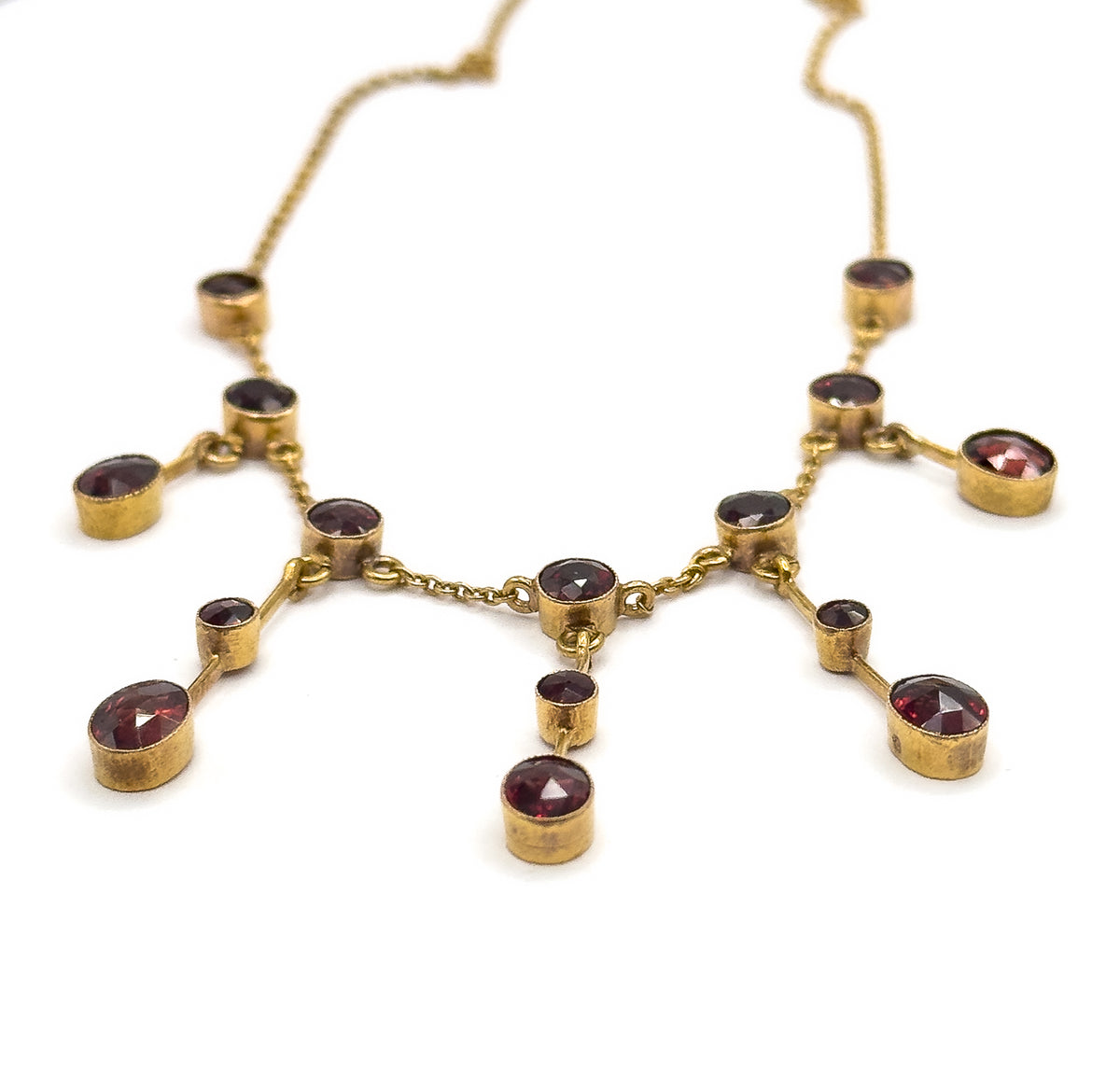 Raised three-quarter view of the whole festoon front, the deep bezel cups tilted so their sides show while the fine chain recedes upward into white, the central garnet drop hanging lowest.