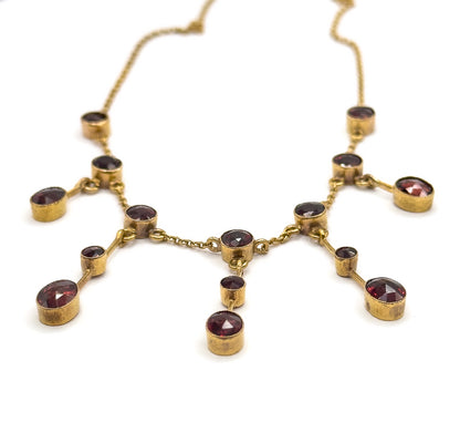 Raised three-quarter view of the whole festoon front, the deep bezel cups tilted so their sides show while the fine chain recedes upward into white, the central garnet drop hanging lowest.