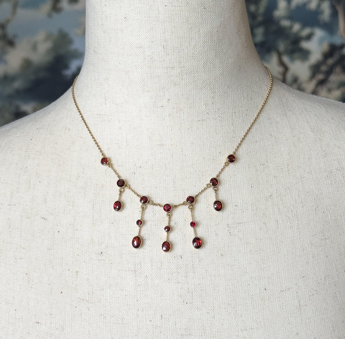 Edwardian garnet festoon necklace in 15K yellow gold, shown at the neck on a linen bust, its bezel-set red stones suspended from fine chain in a draped fringe of small drops.