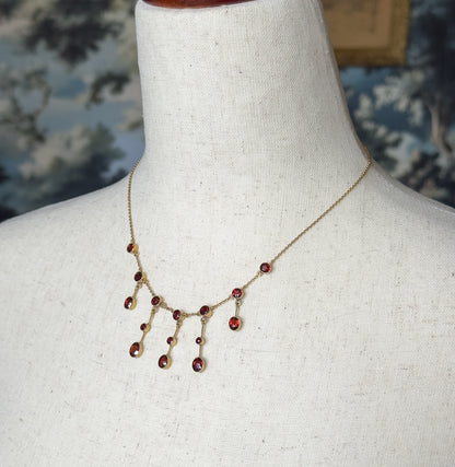 Closer view of the fringe against the linen bust, where the bezel-set garnets hang on short gold bars of differing lengths, with the fine chain curving up to the shoulders past a blue patterned backdrop.