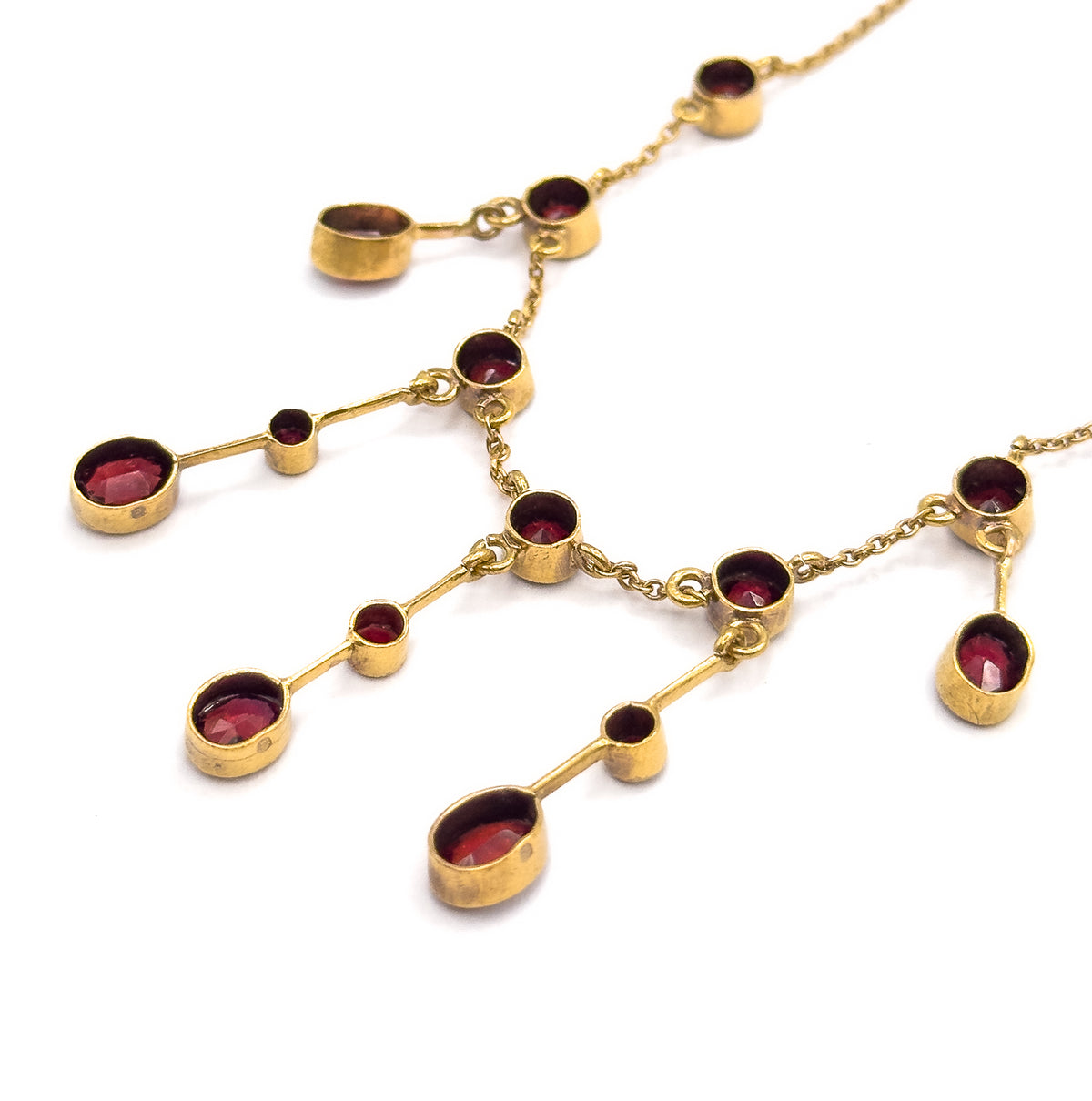 Angled close view laid flat on white, showing the bezel-set garnets and their milgrain frames from the side, with the slim gold bars and chain links joining each drop clearly visible.