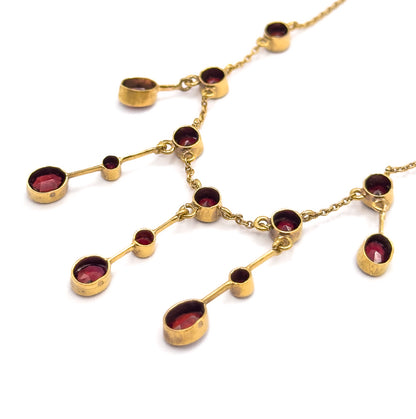 Angled close view laid flat on white, showing the bezel-set garnets and their milgrain frames from the side, with the slim gold bars and chain links joining each drop clearly visible.