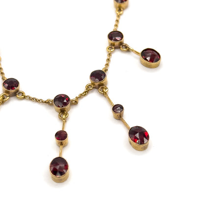 Tight crop on two fringe drops, the milgrain-edged bezels and their jump-ring joins to the cable chain clearly visible, each short gold bar carrying a small garnet above a larger one.