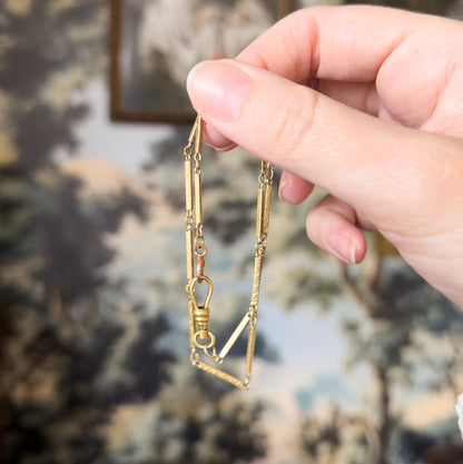 Edwardian ornate bar link watch chain in 14K gold, held draped from fingertips so its slim bar links and dog clip hang free, the hand giving a sense of its small scale.