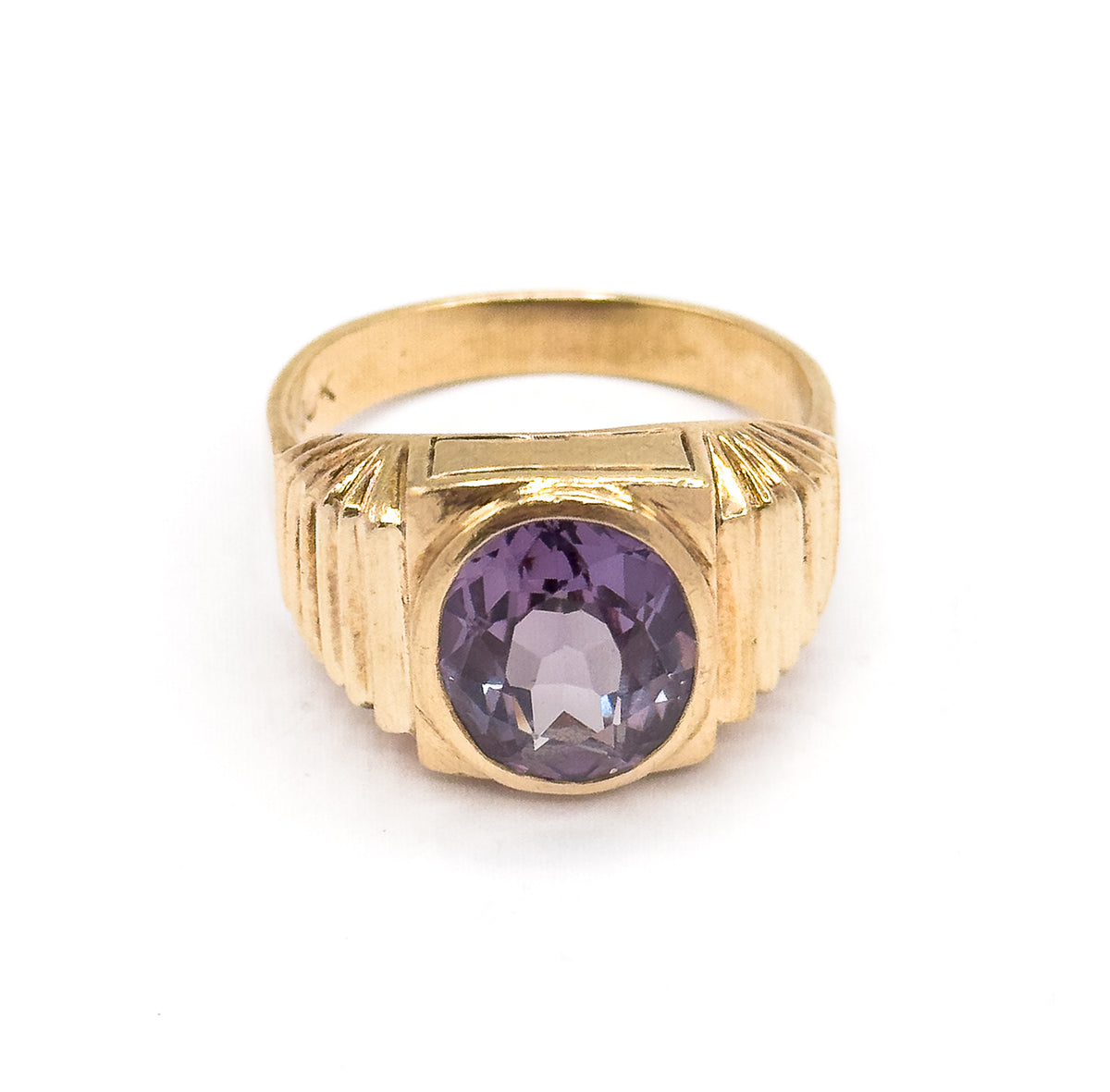 Head-on from the front with the ring standing upright, the oval sapphire violet in its smooth bezel between stepped fluted shoulders, the yellow gold shank arching behind.