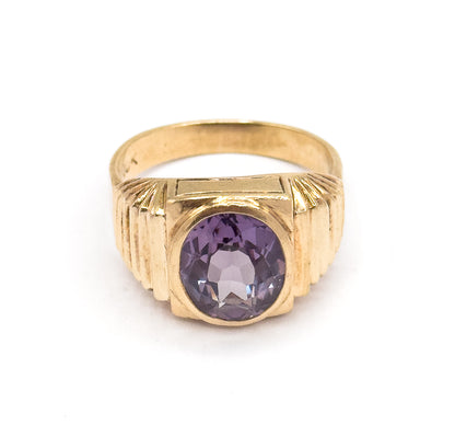 Head-on from the front with the ring standing upright, the oval sapphire violet in its smooth bezel between stepped fluted shoulders, the yellow gold shank arching behind.