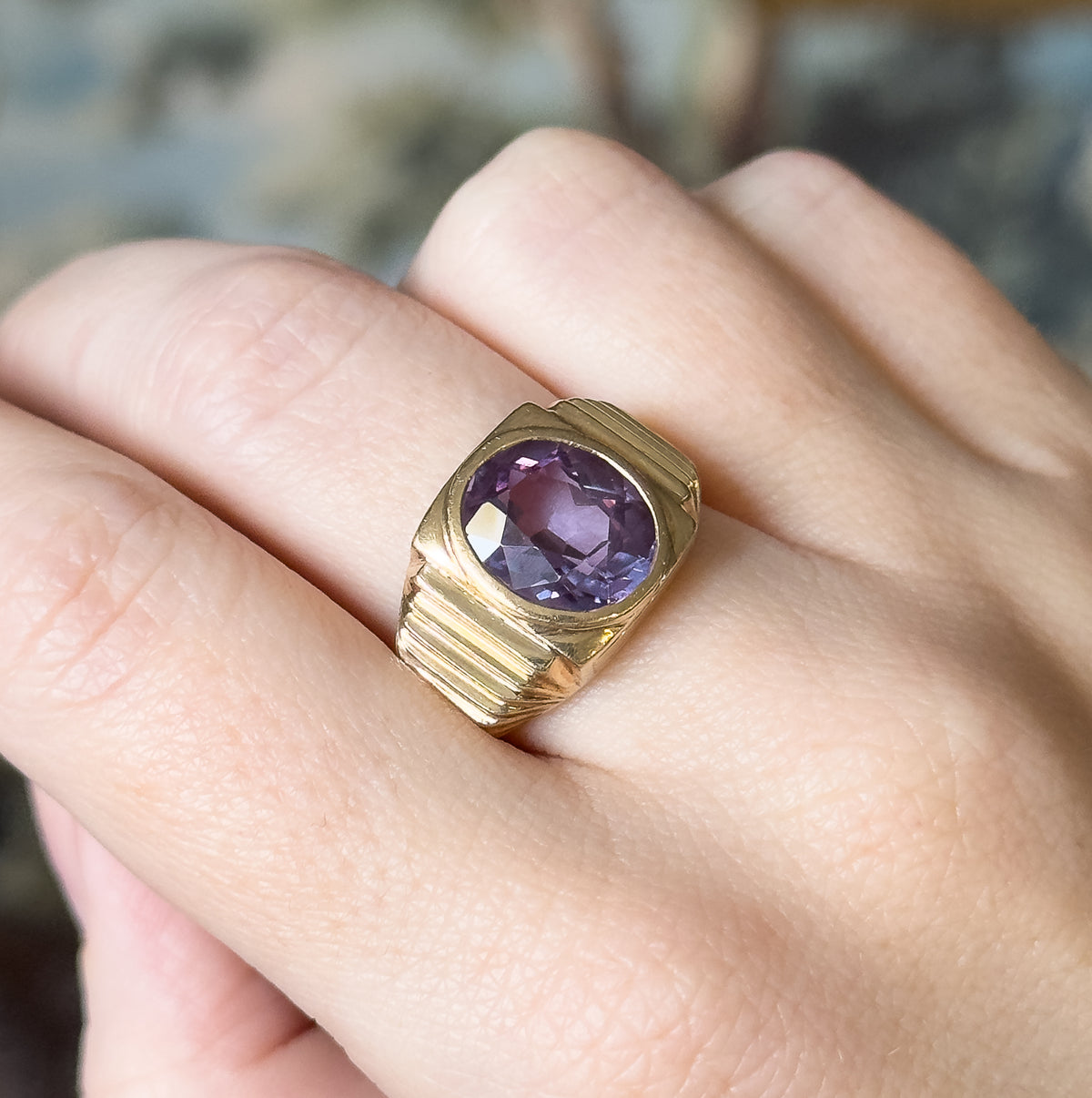 Mid-Century lab-created colour change sapphire ring in 10K yellow gold, shown on a finger, the large oval purple stone in a smooth bezel with ridged shoulders tapering to a wide shank.