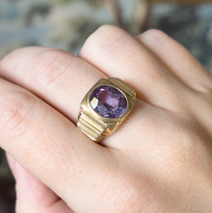 Mid-Century lab-created colour change sapphire ring in 10K yellow gold, shown on a finger, the large oval purple stone in a smooth bezel with ridged shoulders tapering to a wide shank.