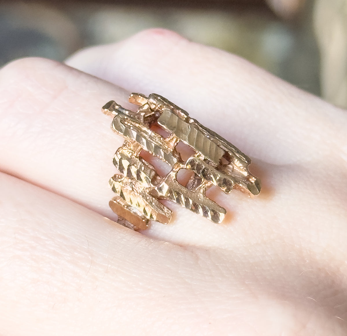 Faceted sculptural ring in 10K yellow gold, shown on a finger in close-up, its open crisscrossing bars of textured metal spanning a wide face that narrows toward the slim shank.