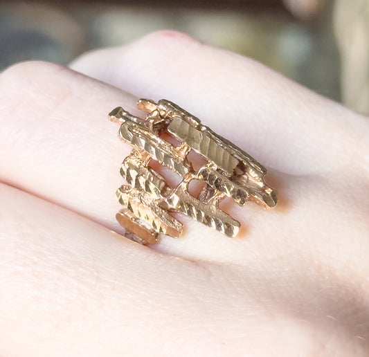 Faceted sculptural ring in 10K yellow gold, shown on a finger in close-up, its open crisscrossing bars of textured metal spanning a wide face that narrows toward the slim shank.