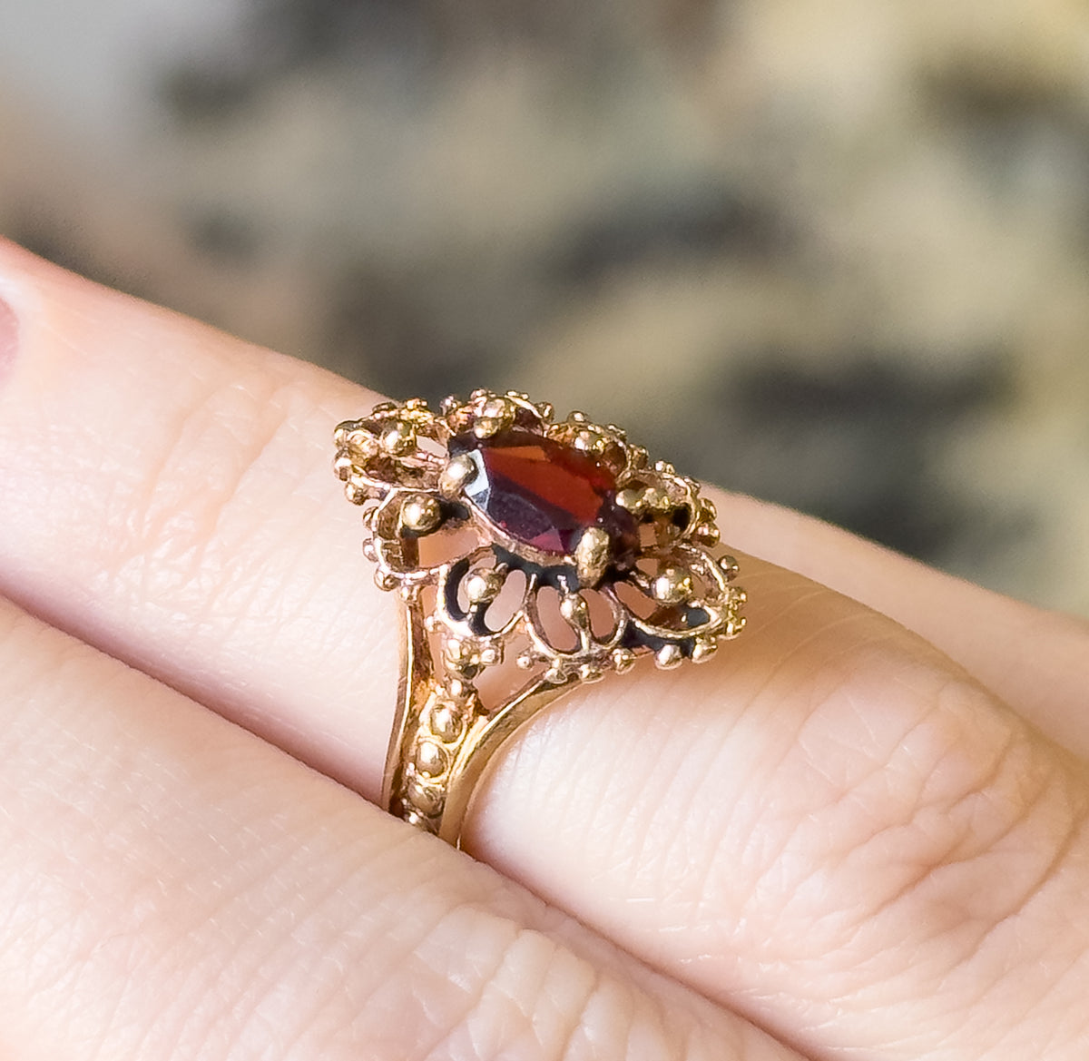 A marquise garnet ring in 10K yellow gold, shown on a finger in close-up, its deep orangish red stone held in a scalloped beaded bezel above a tapering shank.