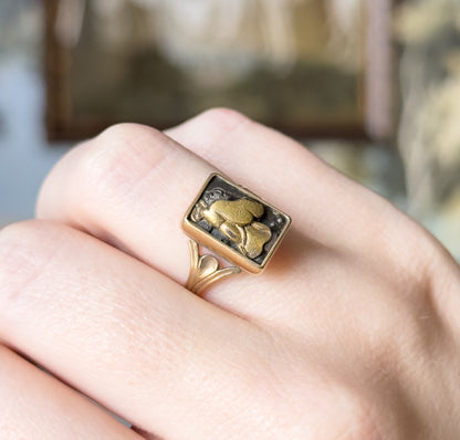 Angled view from above, shown on a finger against a blurred interior, the dark rectangular panel's raised figure catching the light as the bezel meets leaf-like flaring shoulders.