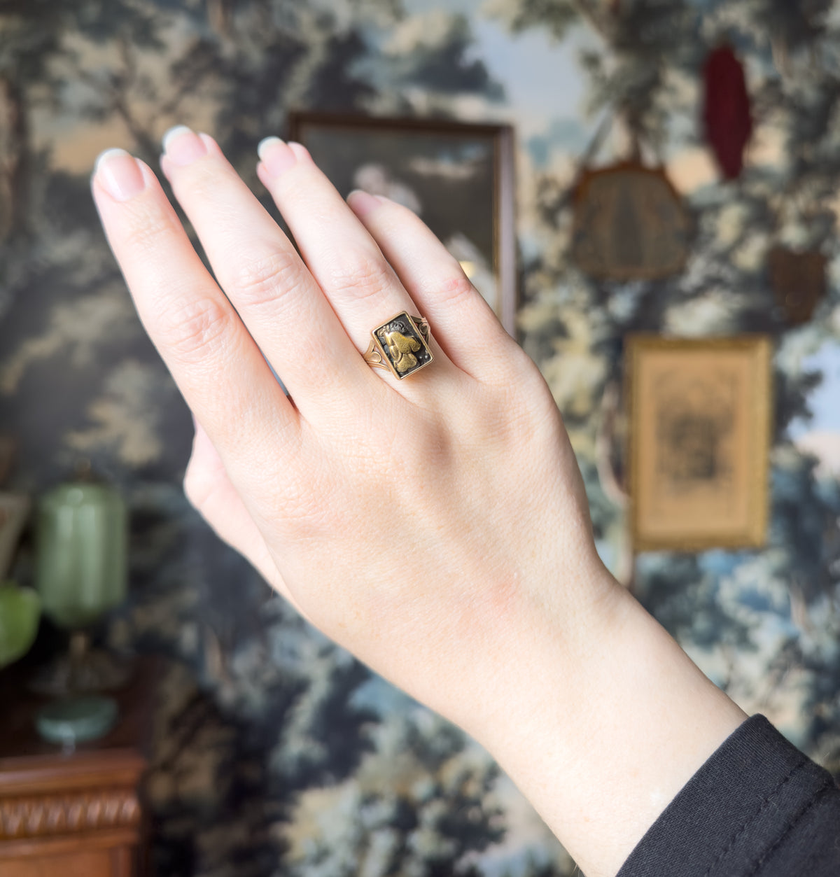 The whole raised hand is in frame against a patterned wall, giving a sense of scale as the small rectangular panel sits on the ring finger beside the unadorned neighbouring fingers.