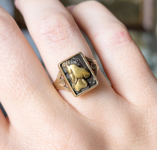 Victorian Shakudo panel ring in 9K yellow gold, shown on a finger, its rectangular dark panel with a raised figural design held in a plain bezel above split, tapering shoulders.