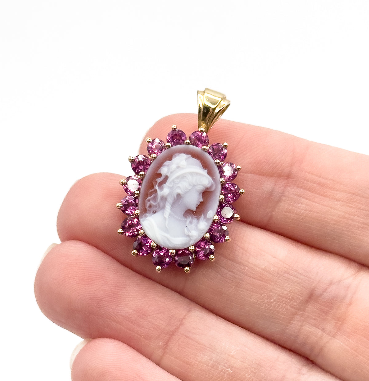 Hardstone cameo and garnet pendant in 14K yellow gold, the carved oval face ringed by prong-set round garnets, shown resting on fingertips to convey its small size.