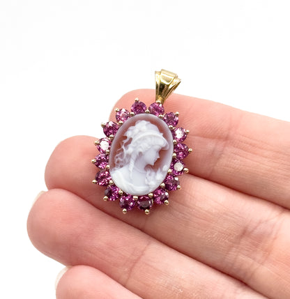 Hardstone cameo and garnet pendant in 14K yellow gold, the carved oval face ringed by prong-set round garnets, shown resting on fingertips to convey its small size.