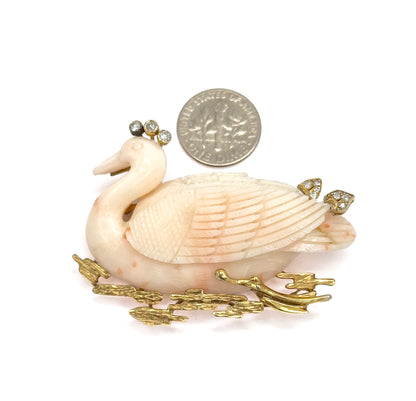 A coin placed beside the brooch for scale, the carved coral swan reaching roughly twice the coin's width, with diamonds at the crown and back and textured gold reeds below.