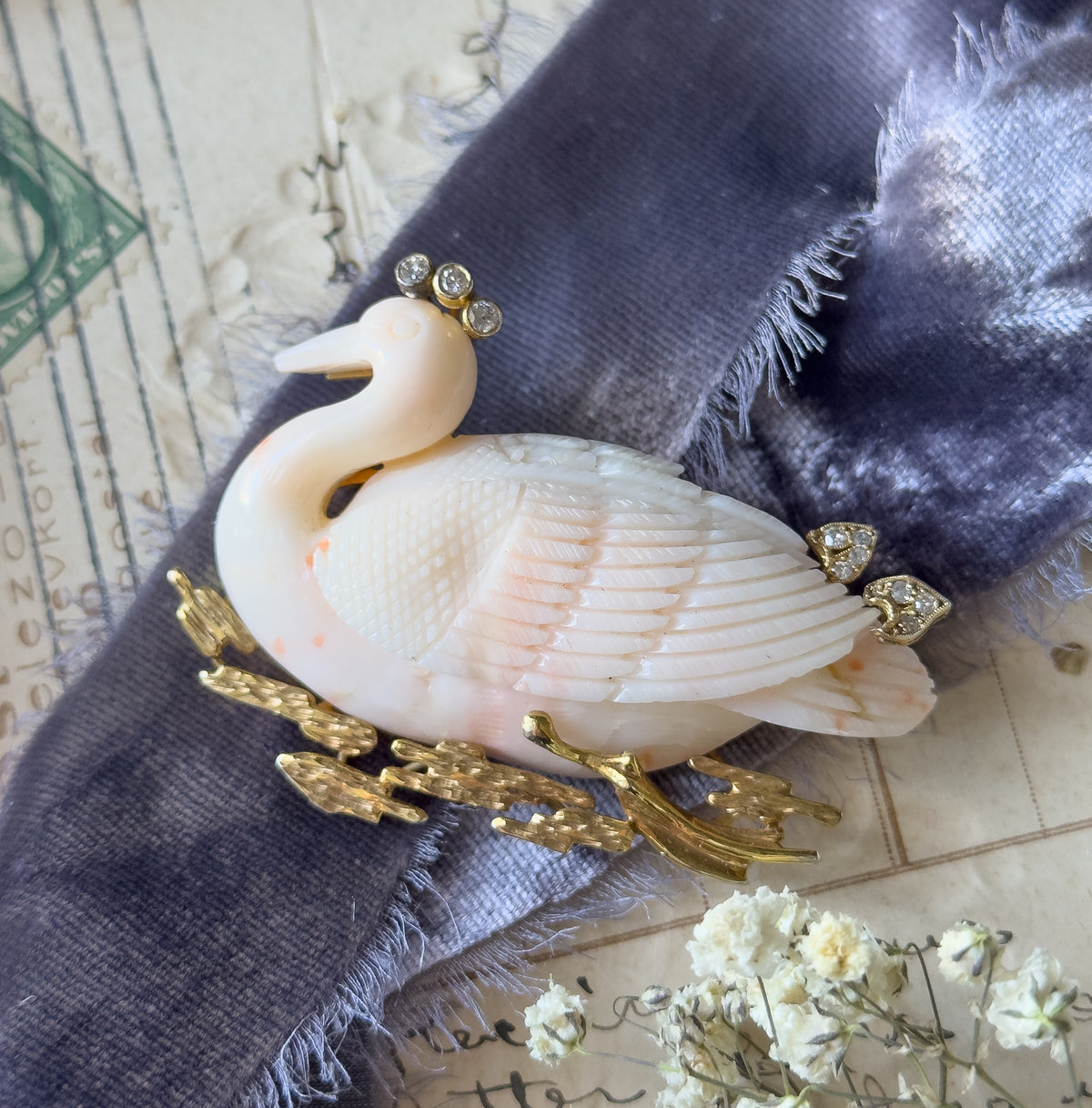 An overhead styled view with the brooch resting on grey velvet ribbon over handwritten paper, dried white flowers at the lower right, the coral swan's carved feathers and diamond accents catching the light.