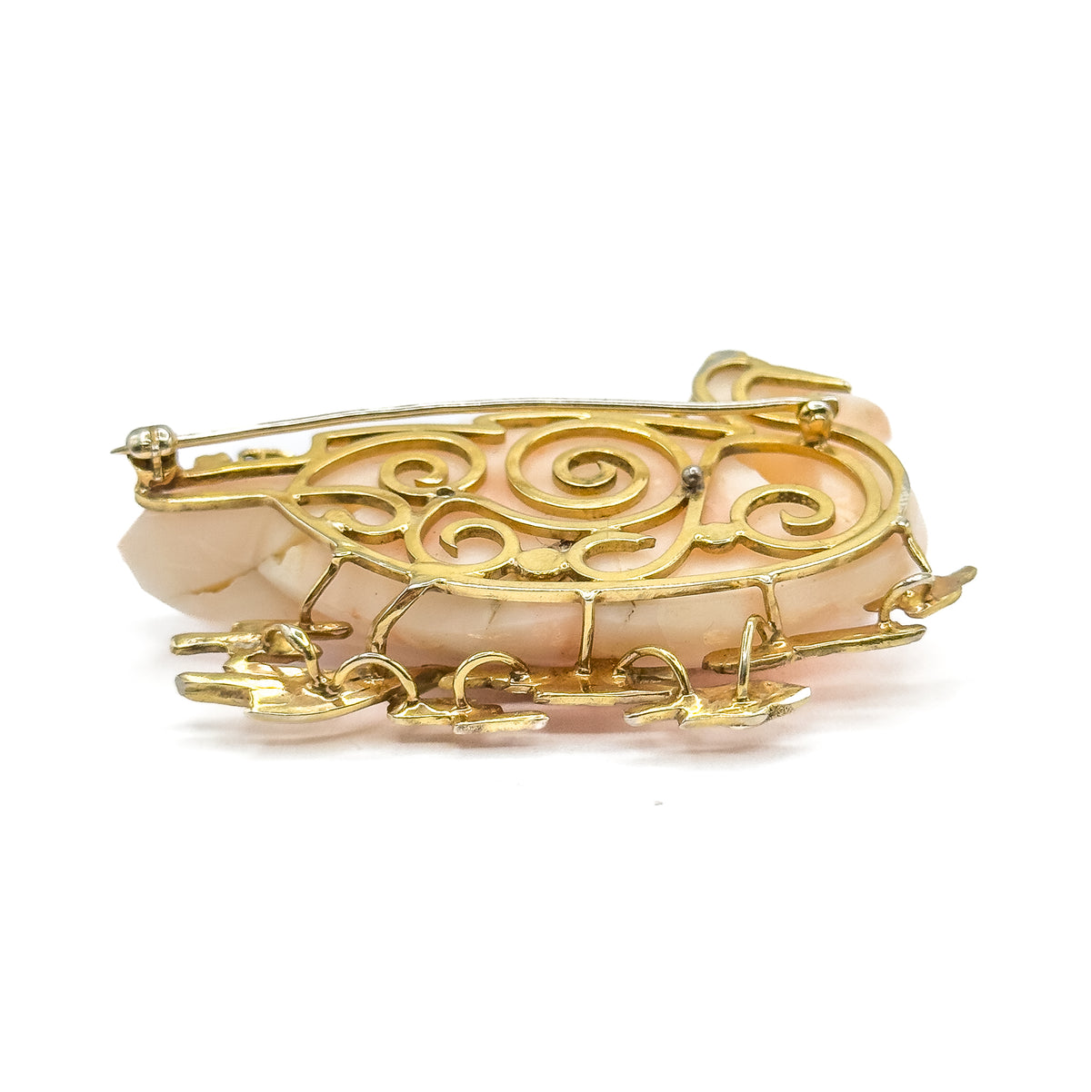 A low side-on view from the back, where the scrollwork gallery curls across the coral's depth and the gold reeds project forward beneath, with the pin stem resting along the upper edge.