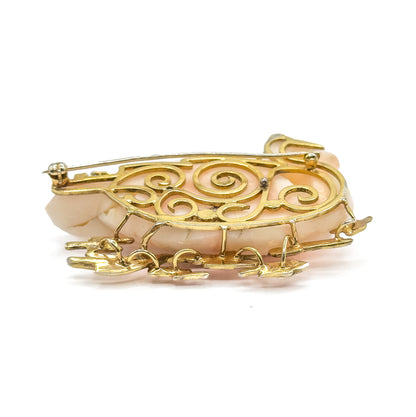A low side-on view from the back, where the scrollwork gallery curls across the coral's depth and the gold reeds project forward beneath, with the pin stem resting along the upper edge.
