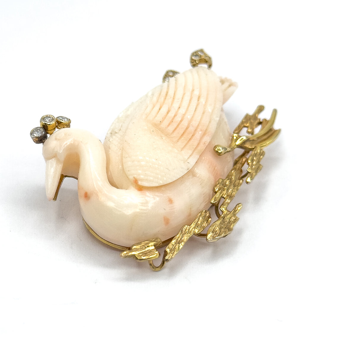 A high three-quarter view from the front, looking down on the carved coral swan's arched neck and feathered back, with the diamond cluster above the head and textured gold reeds fanning out at the side.